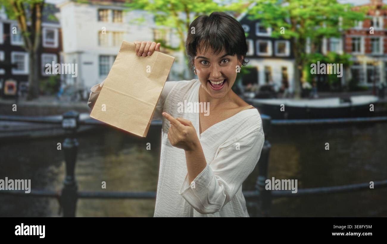 Die Frau zeigt mit dem Finger auf die Papiertüte auf der Kanalstraße, hält sie hoch und lächelt breit; Überraschung. Stockfoto