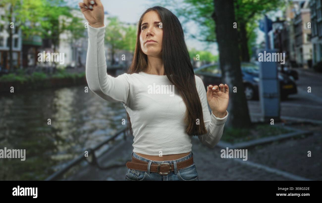 Junge Frau mit weißem Langarmtop und Jeans zeigt mit erhobenen Händen den Finger nach oben neben einem Kanal auf einer gepflasterten Straße. Konzentration. Stockfoto