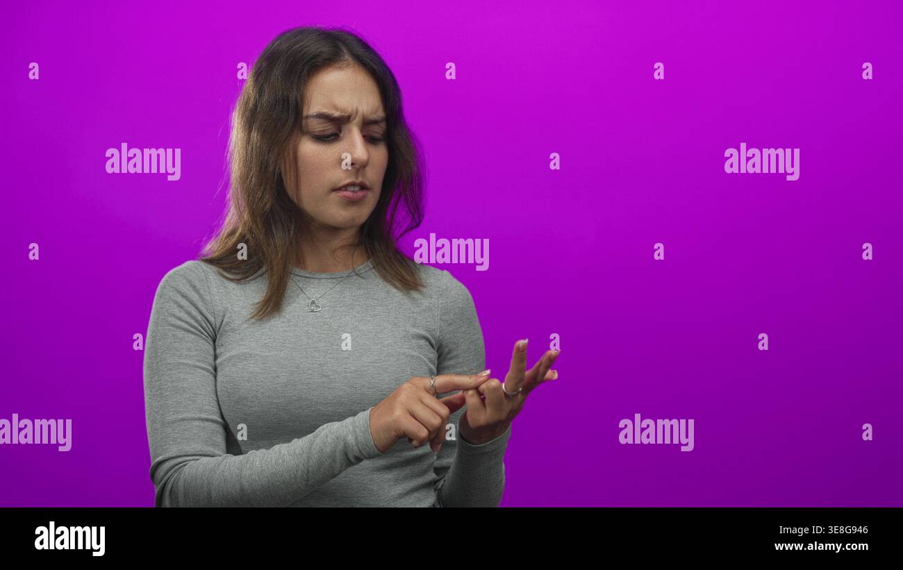 Junge hispanische Frau, die ihre Fingerspitze untersucht, während sie mit der anderen Hand im Studio vor einem violetten Hintergrund zeigt; Sorge. Stockfoto