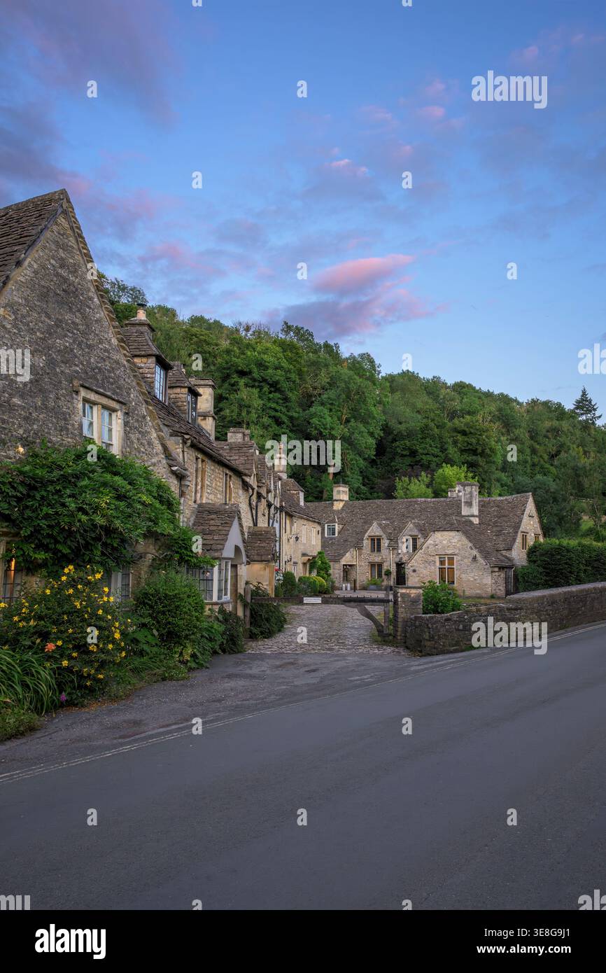 Traditionelle Cotswold Steinhäuser entlang der ruhigen Dorfstraße in Castle Combe, Wiltshire, England bei Sonnenuntergang mit rosa Wolken Stockfoto