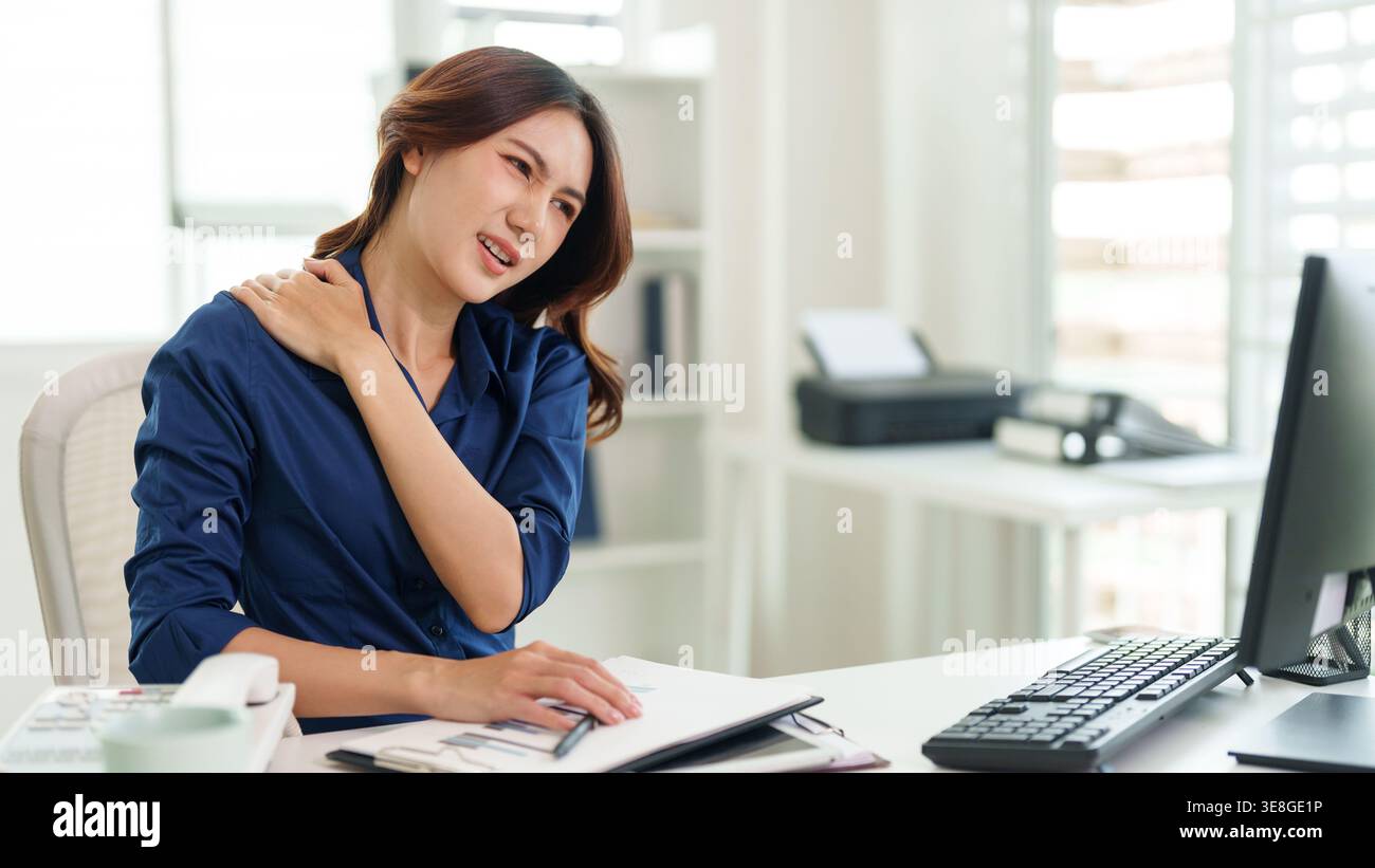 Erwachsene asiatische Frau sitzt am Schreibtisch und hält ihre Schulter schmerzhaft mit einem grimmigen Ausdruck. Die Tastatur des Computermonitors und das Notebook sind auf dem des sichtbar Stockfoto