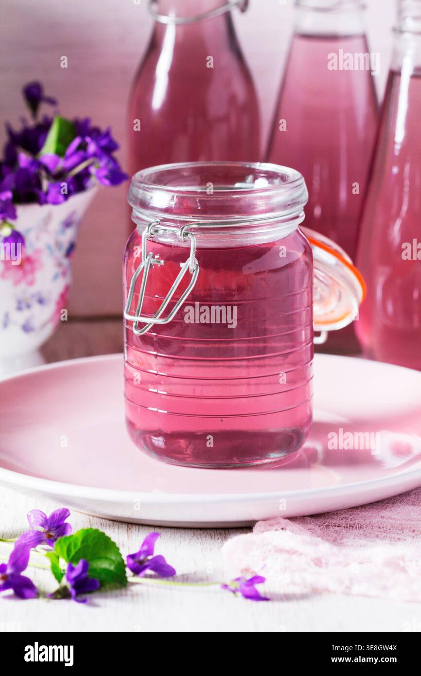 Violetter Sirup in einem Glasgefäß, kombiniert mit einem Bouquet frischer Veilchen, kreiert eine zarte florale Komposition mit weichen Pastelltönen und natürlichem Licht. Stockfoto