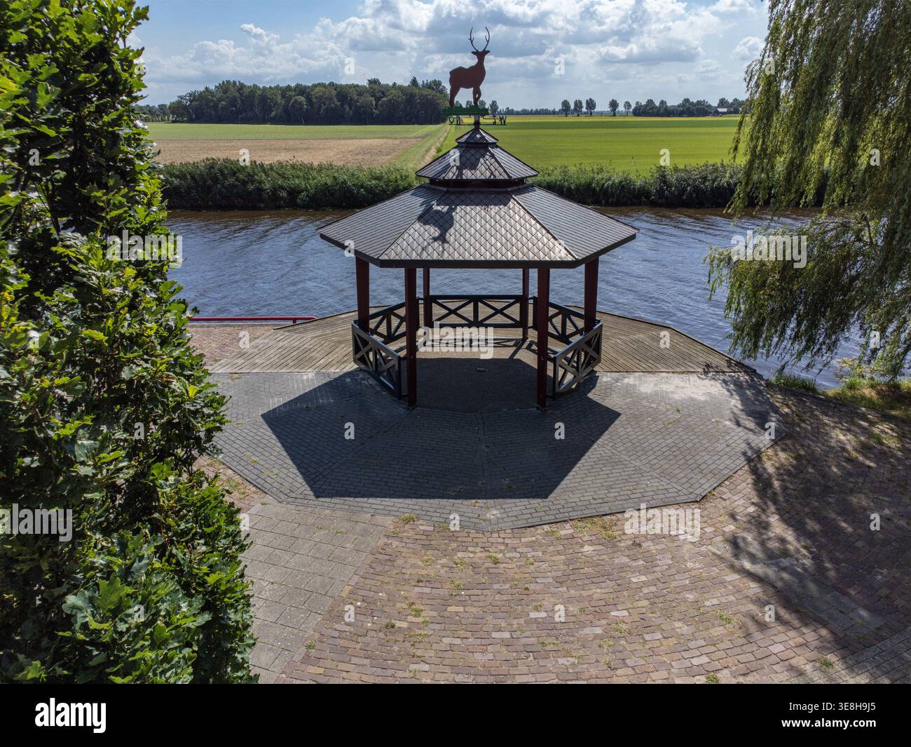 Blick aus der Vogelperspektive auf einen Pavillon mit einer Hirschstatue, stolz vor einer Kulisse aus grünen Feldern und einem ruhigen Kanal, Tollebeek, Flevoland, Net Stockfoto