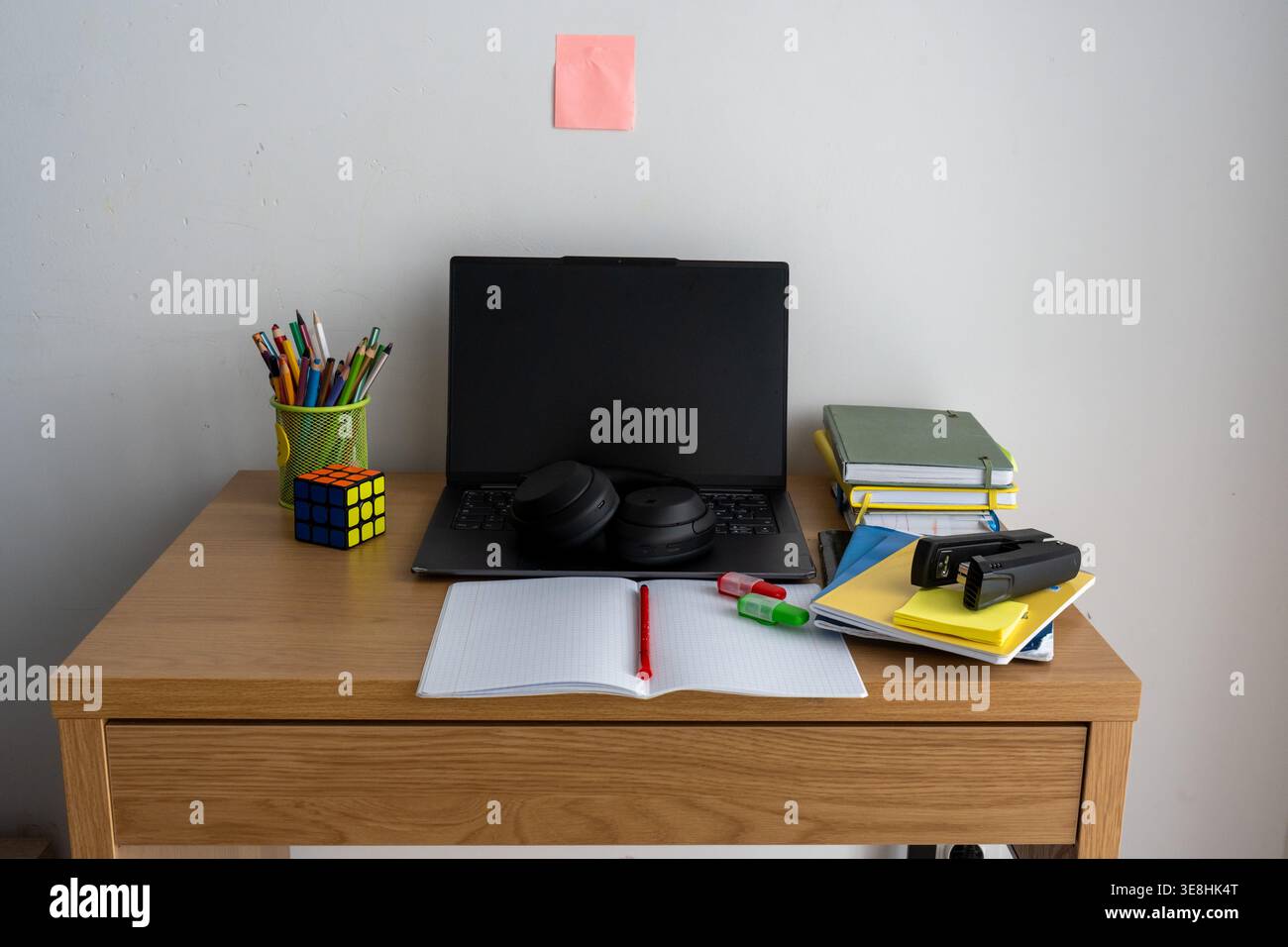 Schüler-Holzschreibtisch mit offenem Laptop, Kopfhörer, bunten Bleistiften, Notizbüchern, Textmarkern, Rubiks Würfel und Haftnotiz an der Wand. Moderner Arbeitsraum Stockfoto