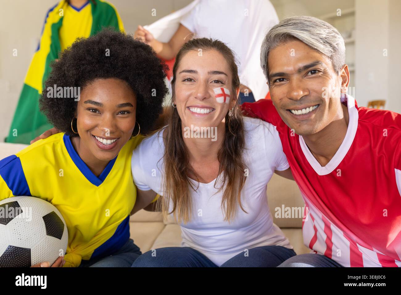 Freunde, die Mannschaftstrikots tragen, jubeln und nach dem Tor auf der Couch posieren und Fußball halten Stockfoto