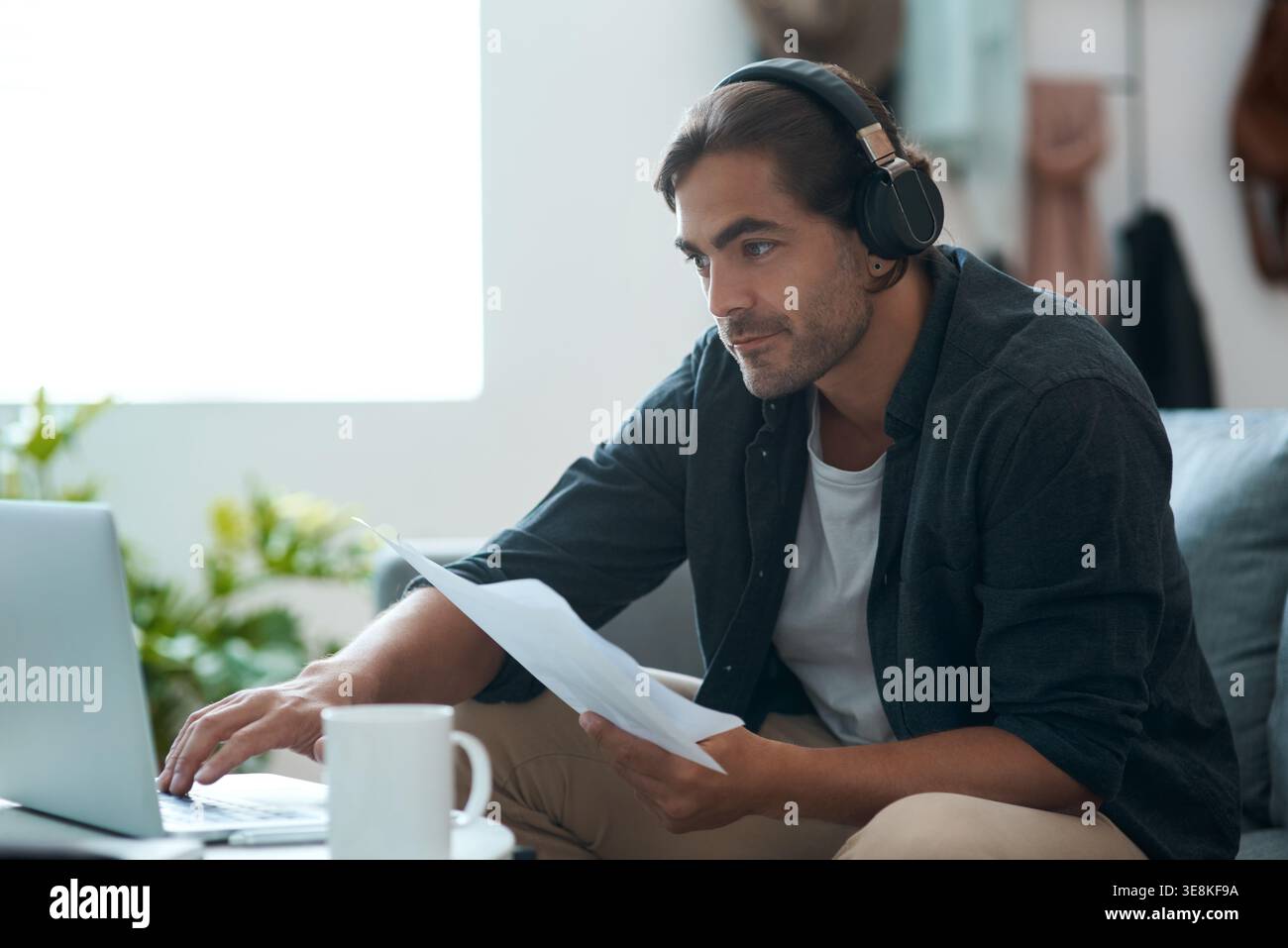 Mann, Laptop und Papierkram mit Fernarbeit von zu Hause aus mit Kopfhörern, Verwaltung und Bewertung auf dem Sofa. Person, virtueller Assistent und Dokumente mit Tippfunktion Stockfoto