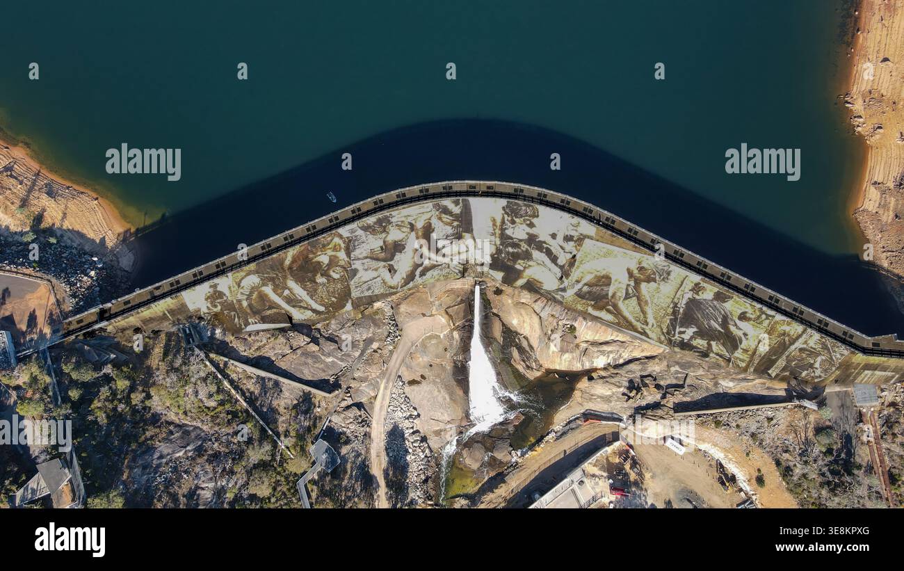 Ein dramatischer Blick von oben nach unten auf einen gebogenen Betondamm, der tiefes blaublaues Wasser zurückhält. Die Dammmauer ist mit einem riesigen sepiafarbenen Wandgemälde bedeckt, das Menschen darstellt Stockfoto