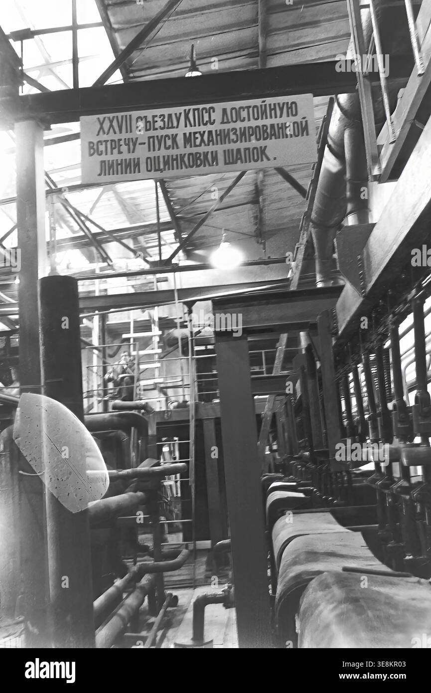 Slawjansk, 1980er Jahre, Werkstatt der Armature and Isolator Plant (AIZ). Das Schwarzweiß-Archivfoto zeigt das industrielle Innenleben des Unternehmens und einen Abschnitt der mechanisierten Verzinkungslinie. Der Kamerawinkel betont das komplexe Rohrleitungssystem, die Aufhängungsmechanismen und die tragenden Träger. Ein Poster zu Ehren des 27. KPdSU-Kongresses ist oben angebracht. Tief in der Werkstatt kann ein ruhender Arbeiter gesehen werden, der dem Leben Authentizität verleiht. Ein echtes Dokument der Epoche, das den friedlichen Donbass zeigt. Stockfoto