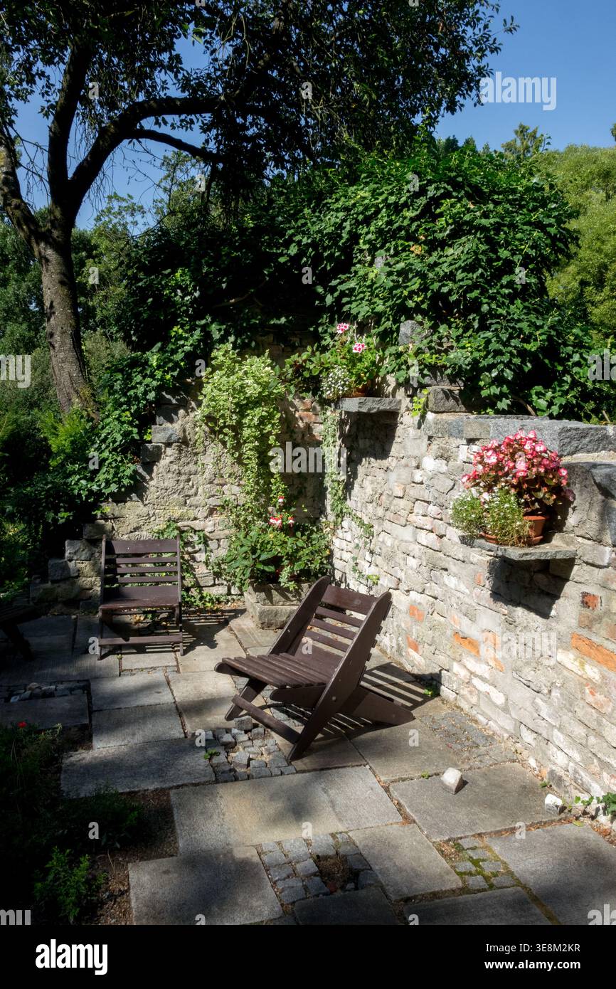Holzstühle auf einer Steinterrasse in einem sonnigen Sommergarten mit Blumen und Steinmauer Stockfoto