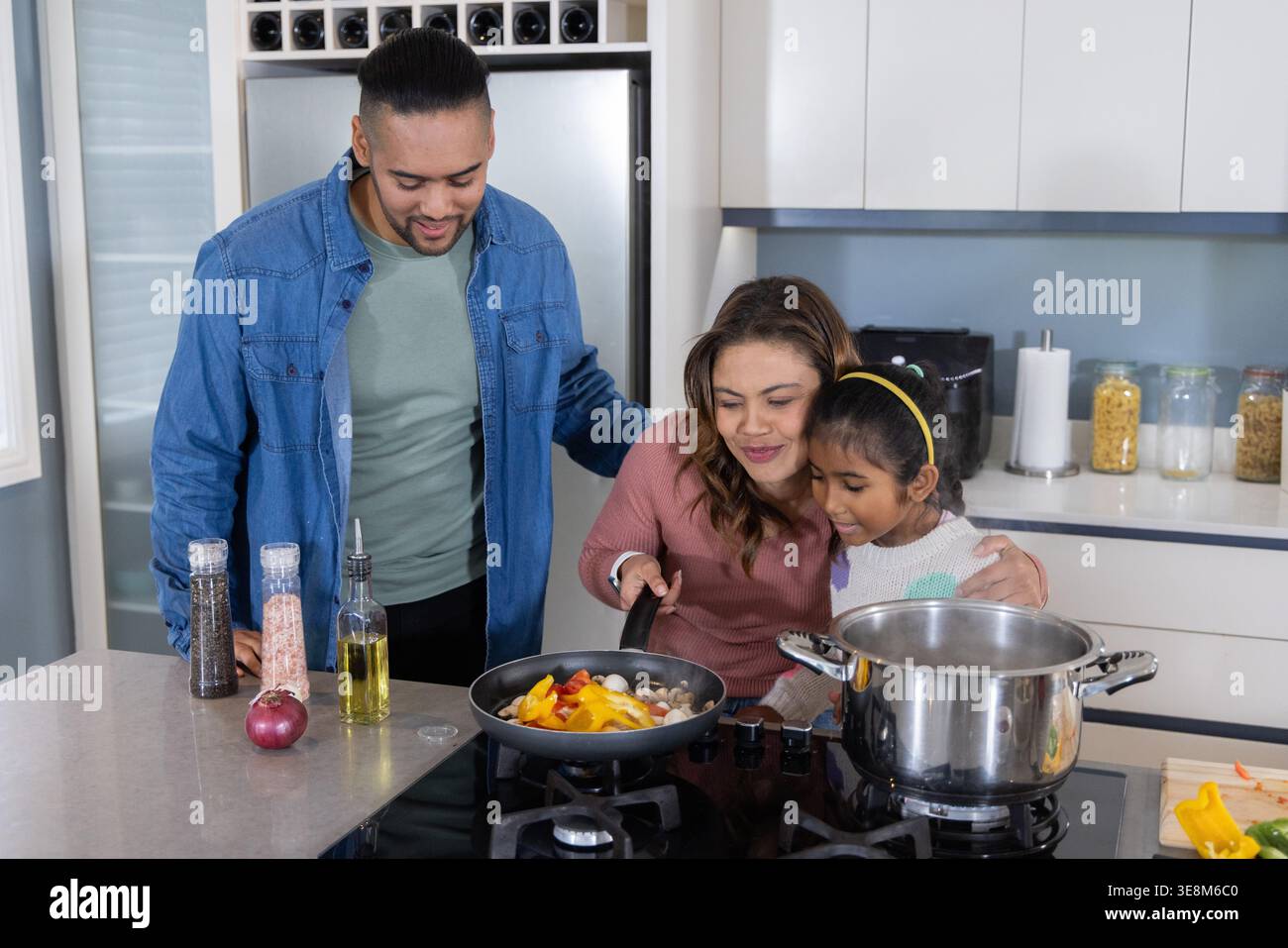 Familieneltern und Tochter kochen Paprika und Pilze in der Pfanne auf dem Kücheninselofen Stockfoto
