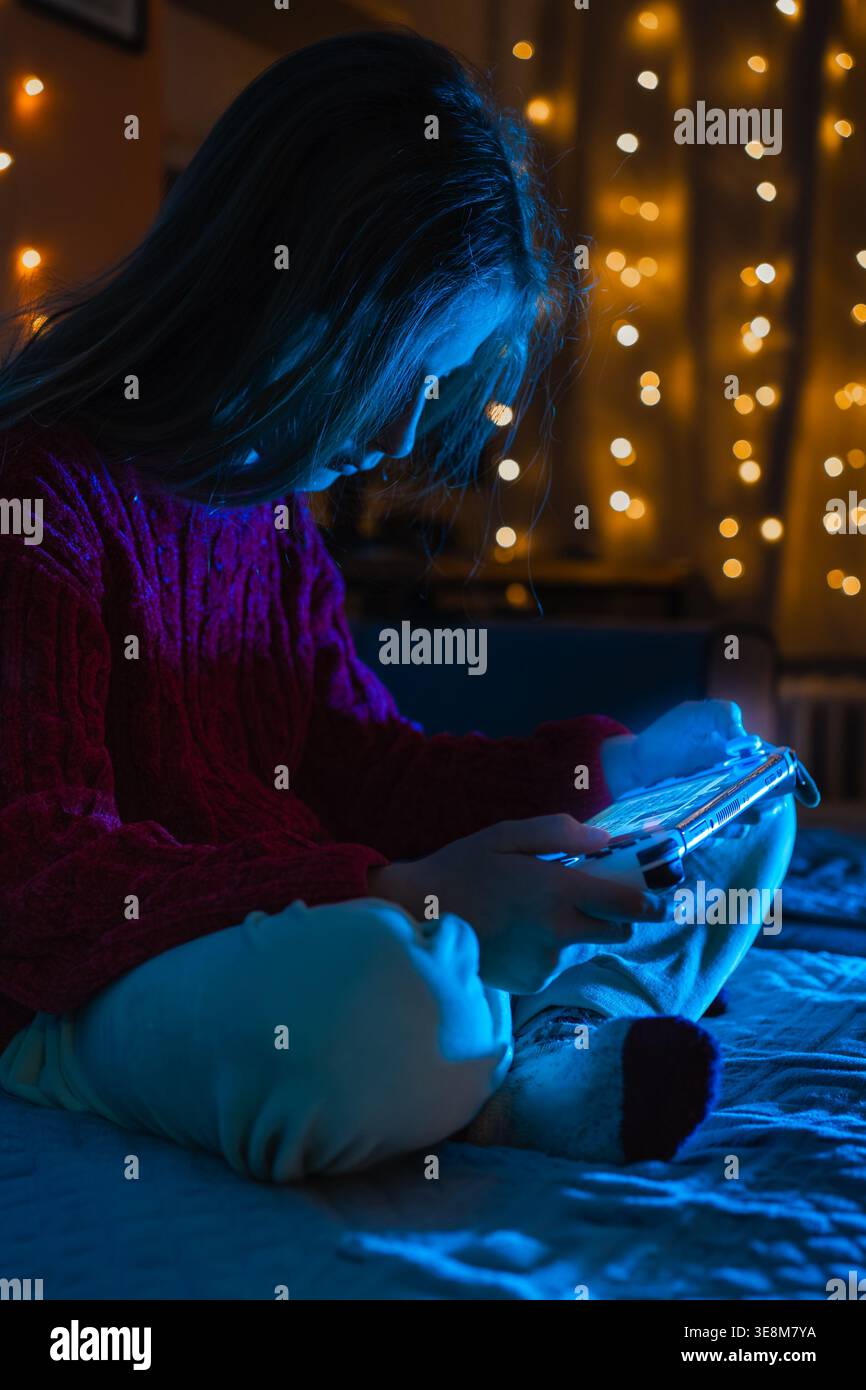 Vertikales Foto eines Mädchens in einem roten Pullover, das auf einer Handkonsole spielt, die von rosa und blauem Licht beleuchtet wird. Stockfoto