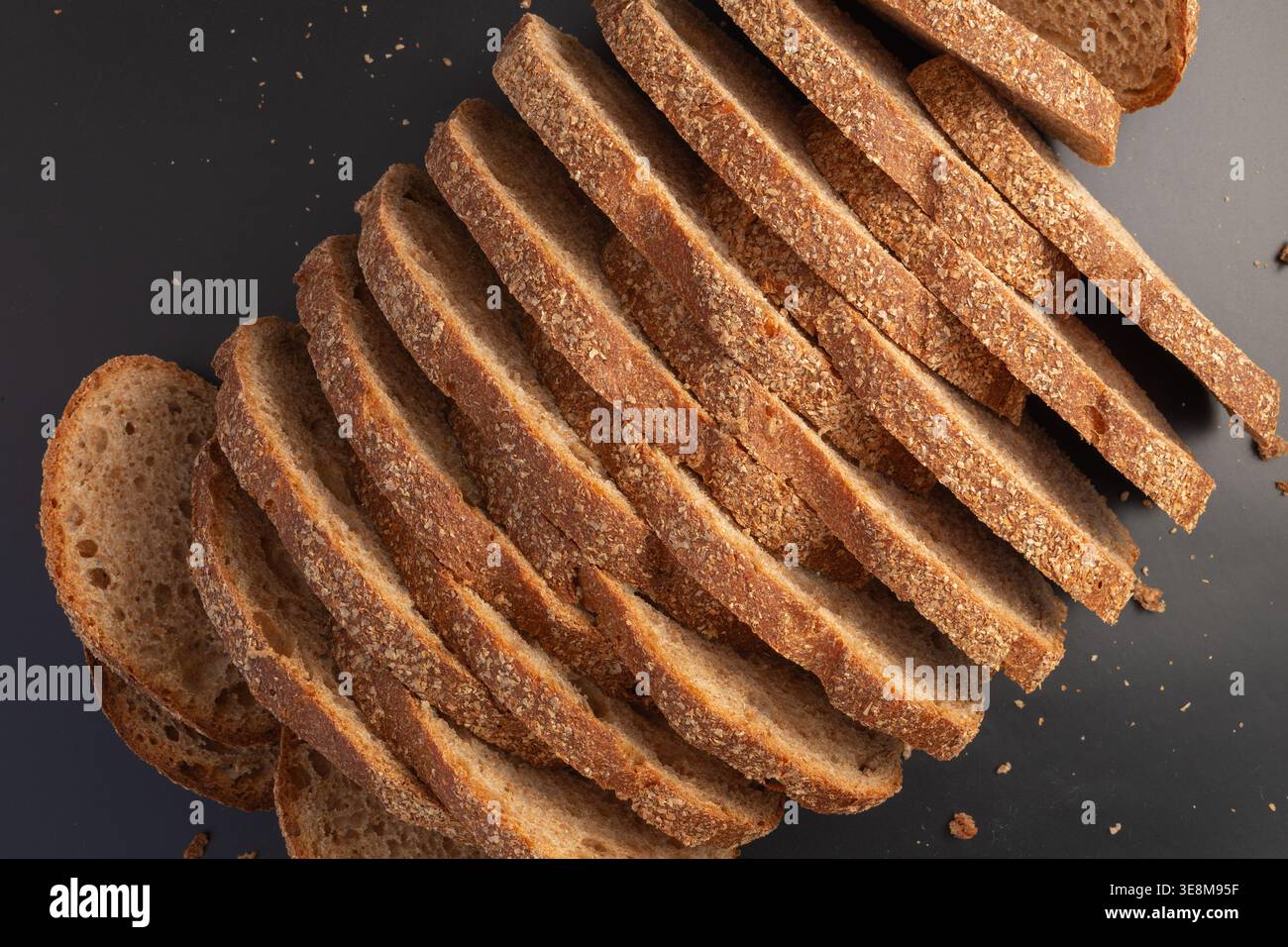 Diagonale Zusammensetzung rustikaler Roggenbrotscheiben auf dunkel matter Oberfläche. Geschnittener Brotlaib aus hellem Roggenbrot auf dunklem Hintergrund Stockfoto