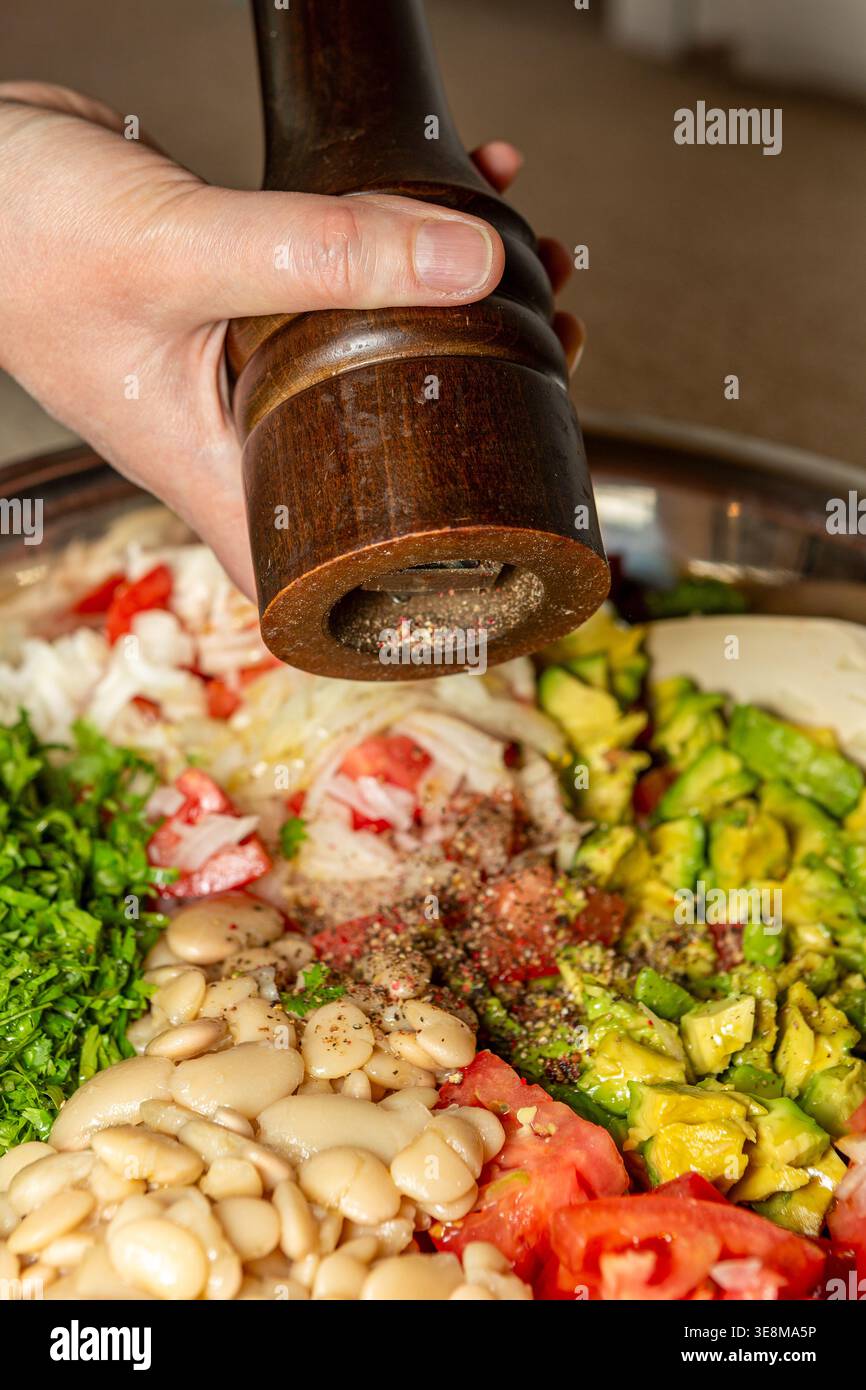 Halten Sie die hölzerne Pfeffermühle von der Hand oben, direkt über dem Salat in der Schüssel, und stauben Sie die Salatzutaten darunter – Avocadostücke, Tomaten, Bohnen. Stockfoto