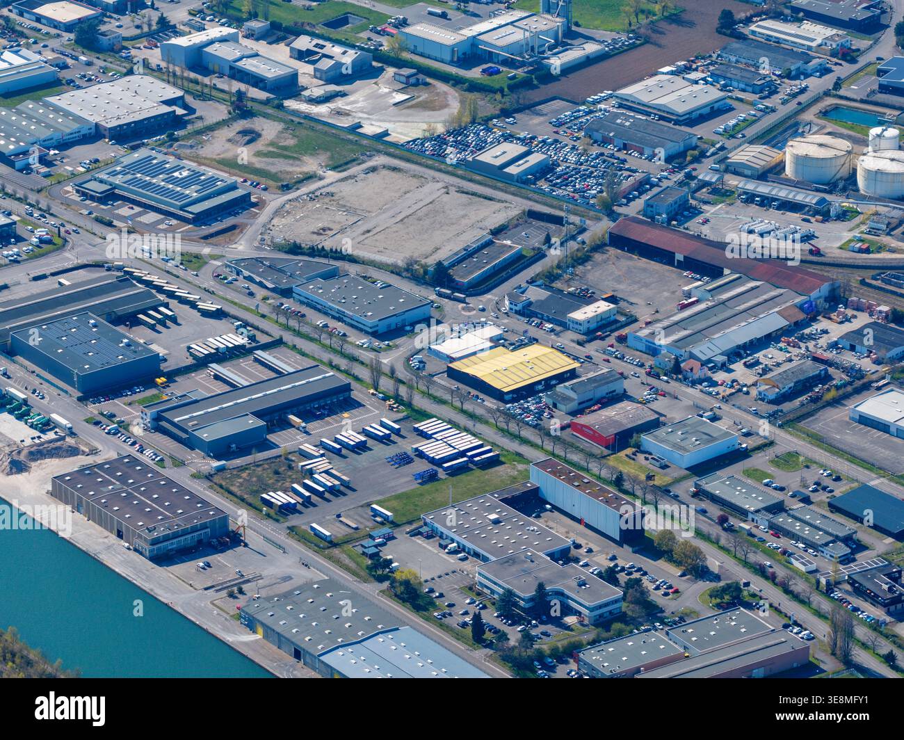 Aus der Vogelperspektive eines weitläufigen Industriegebiets, in dem sich die starke Geometrie der Lagerhäuser und Fabriken mit dem sich schlängelnden Kanal Sausheim, Grand, abhebt Stockfoto