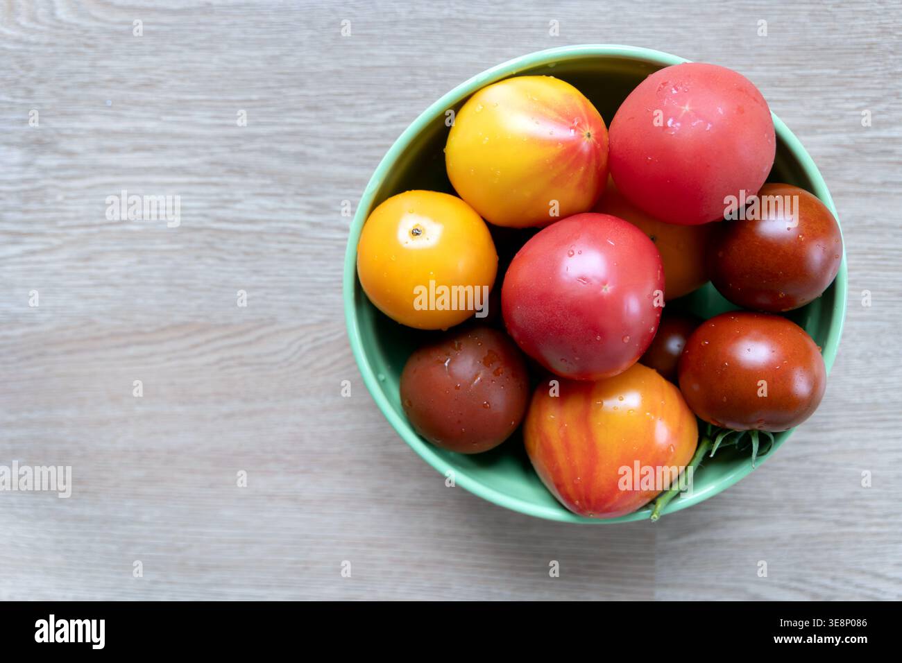 Verschiedene Tomaten in einer grünen Schüssel auf Holzoberfläche. Verschiedene frische Tomaten, darunter rote, gelbe und braune Tomaten, werden in eine grüne Schüssel auf einem gelegt Stockfoto