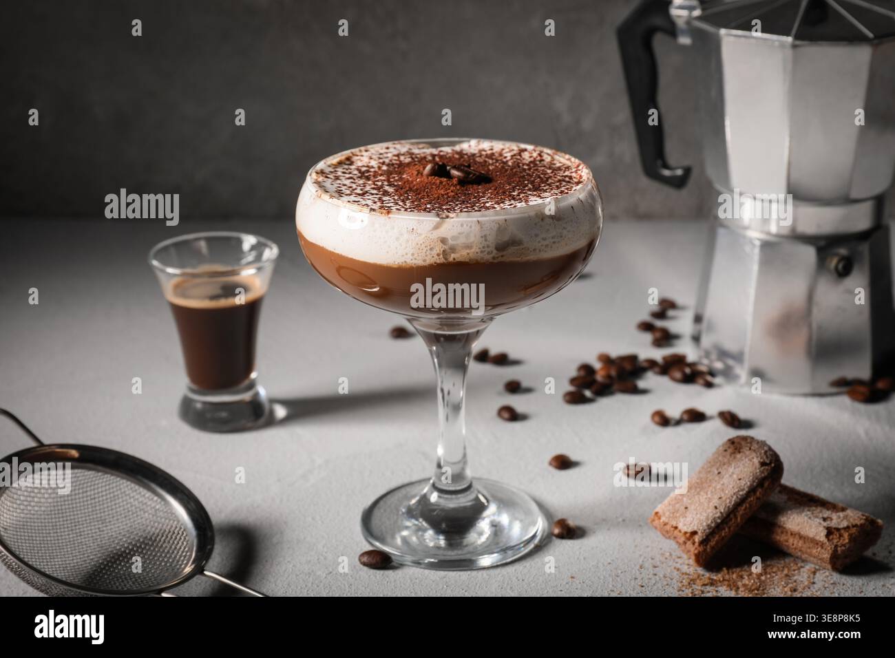 Glas leckerer Tiramisu Cocktail mit Geysir-Kaffeemaschine, Bohnen, Savoiardi und Sieb auf grauem Hintergrund Stockfoto