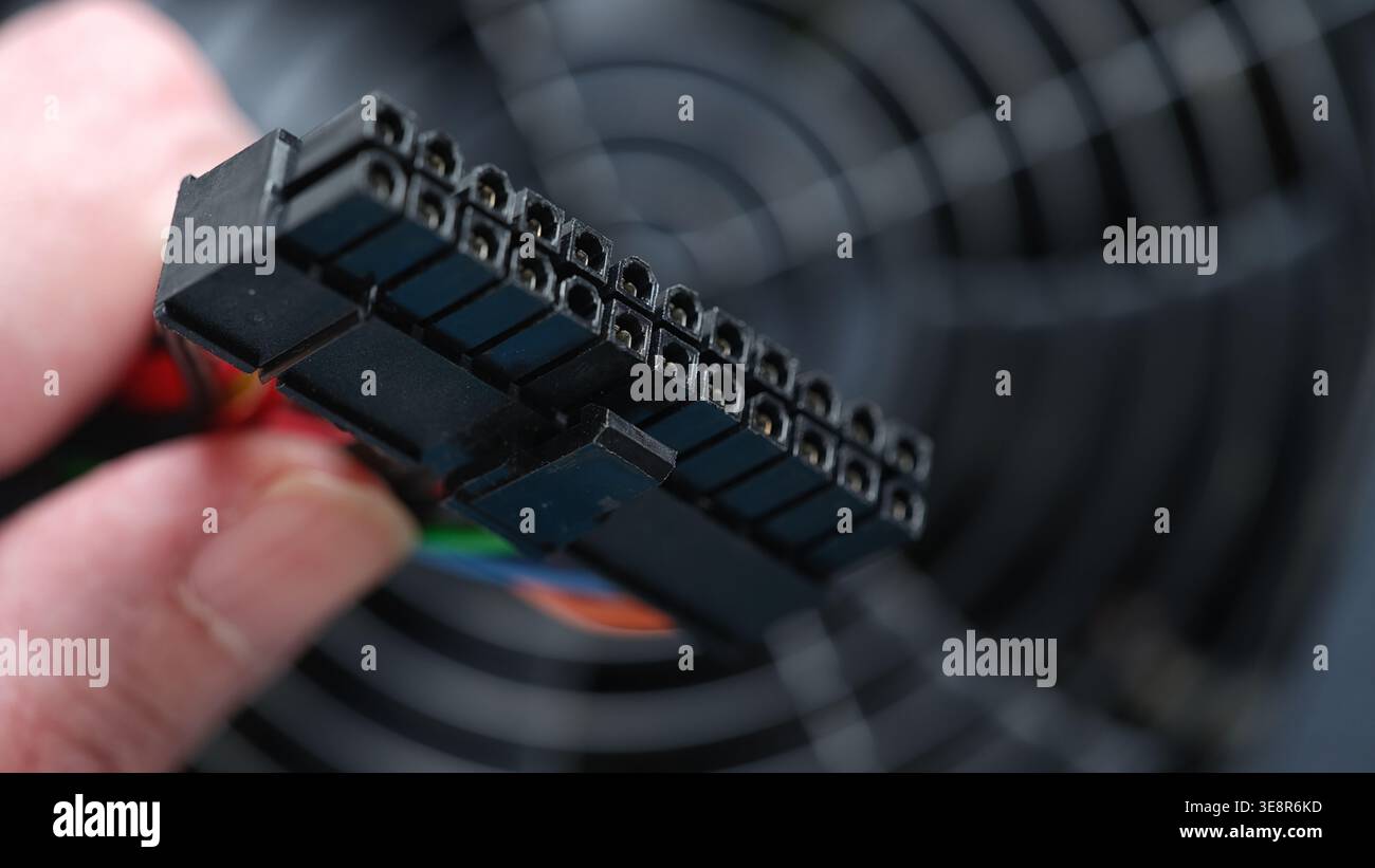 Eine Hand hält einen 24-poligen ATX-Netzstecker mit sichtbarem Staub auf seiner Oberfläche gegen das Lüftergitter eines Computer-Netzteils. Nahaufnahme. Stockfoto