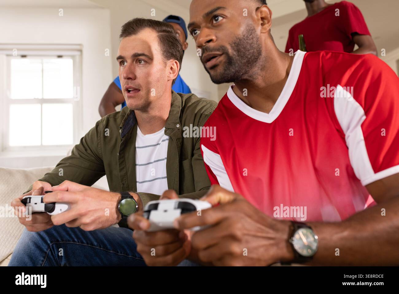 Verschiedene männliche Freunde lehnen sich nach vorne und spielen Konsolenspiel auf einem hellen Sofa mit Controllern Stockfoto