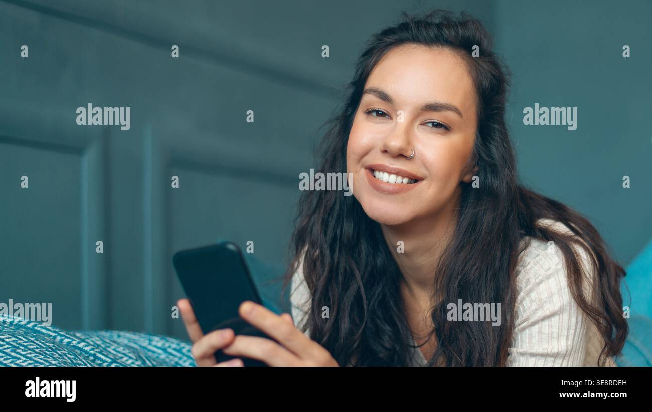 Frau lächelt tagsüber in die Kamera, während sie das Smartphone in gemütlicher Innenumgebung nutzt Stockfoto