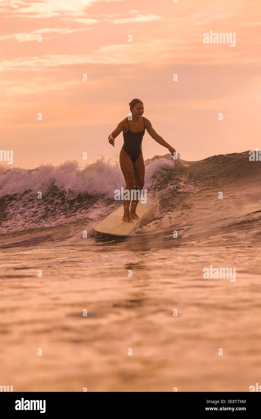 Frau Tanzt Auf Surfbrett Unter Sonnenuntergang, Arme In Celebration Erhoben, Während Sie Sanfte Schwellen Reiten, Verspielter Rhythmus, Stockfoto
