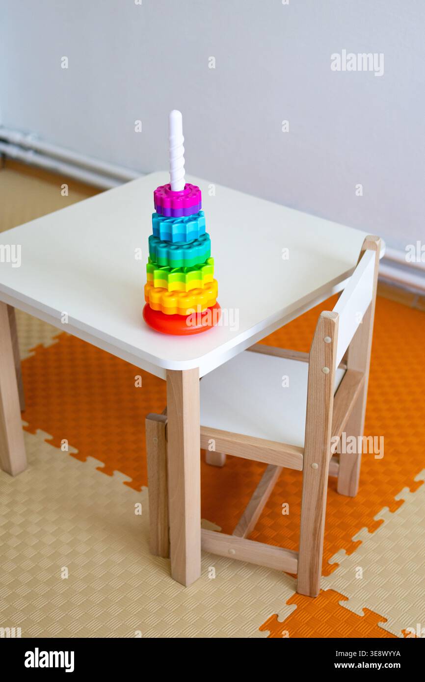 Kindergartenmöbel mit einem Holzstuhl neben einem weißen Tisch, auf dem ein farbenfrohes Spielzeugstapel steht. Helle Schaumstoffmatten bilden ein Orange Stockfoto