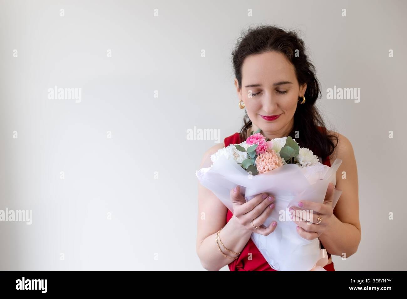 Frau in rotem Kleid genießt einen Blumenstrauß, lächelt mit geschlossenen Augen und hält frische Blüten. Leerzeichen für Text. Stockfoto