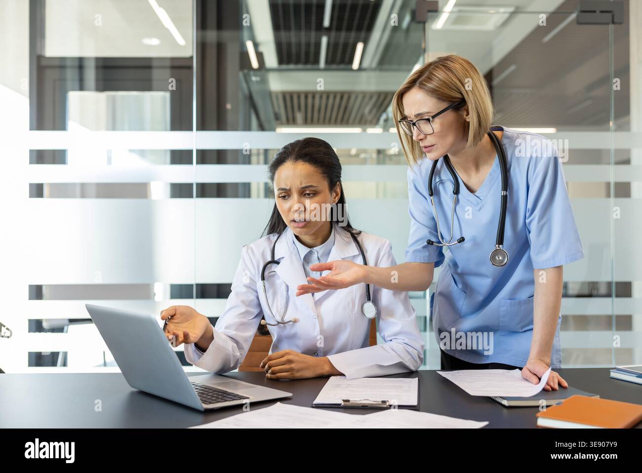Zwei Ärztinnen arbeiten zusammen und analysieren medizinische Dokumente und Informationen auf einem Laptop während eines ernsthaften Gesprächs in einem modernen Büro oder einer Klinik Stockfoto