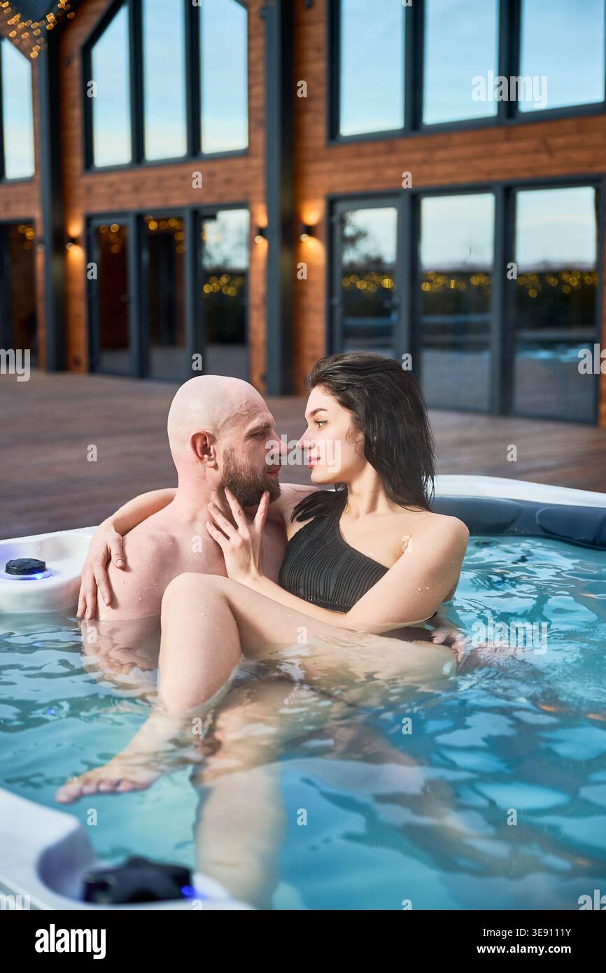 Paare sitzen im Whirlpool und schauen sich gegenseitig an. Mann und Frau genießen intime Spa-Momente im Freien in der Nähe eines modernen Hauses, Romantik, Wellness, Luxus-Lifestyle-Konzept Stockfoto