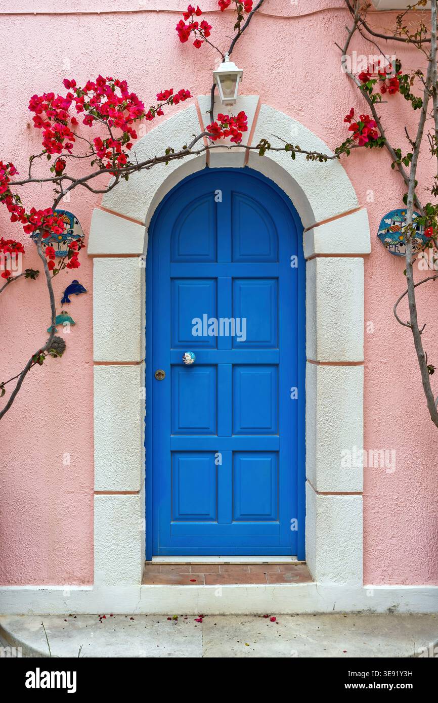 Hellblaue bogenförmige Holztür in einer pinkfarbenen Stuckwand mit weißem Steinrahmen, verziert mit Bougainvillea-Blumen und Keramikschmuck. Stockfoto