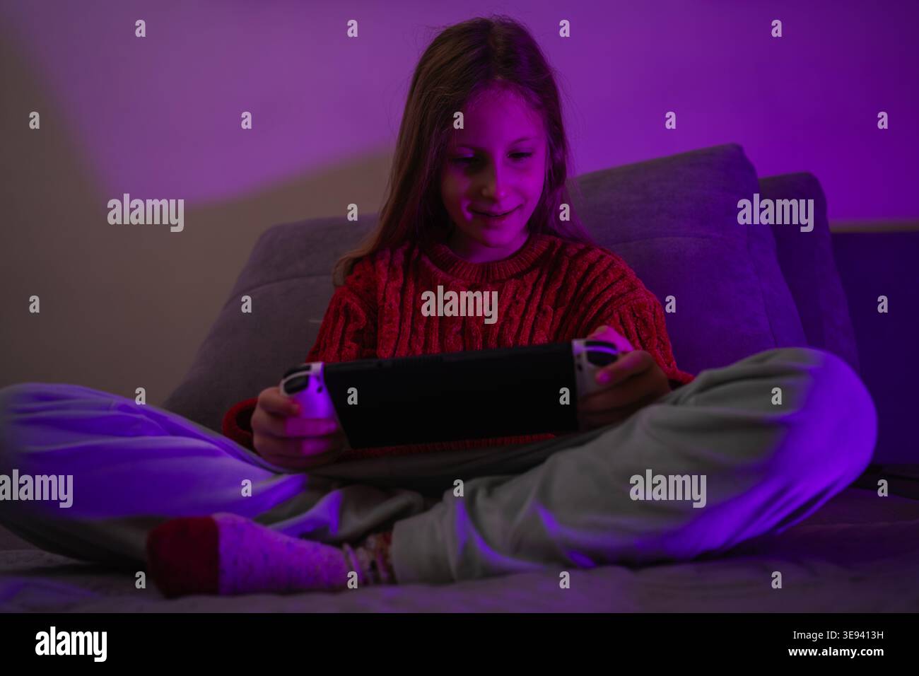 Lächelndes Mädchen, das ein Videospiel auf einem tragbaren Gerät spielt, das von violettem Umgebungslicht beleuchtet wird Stockfoto