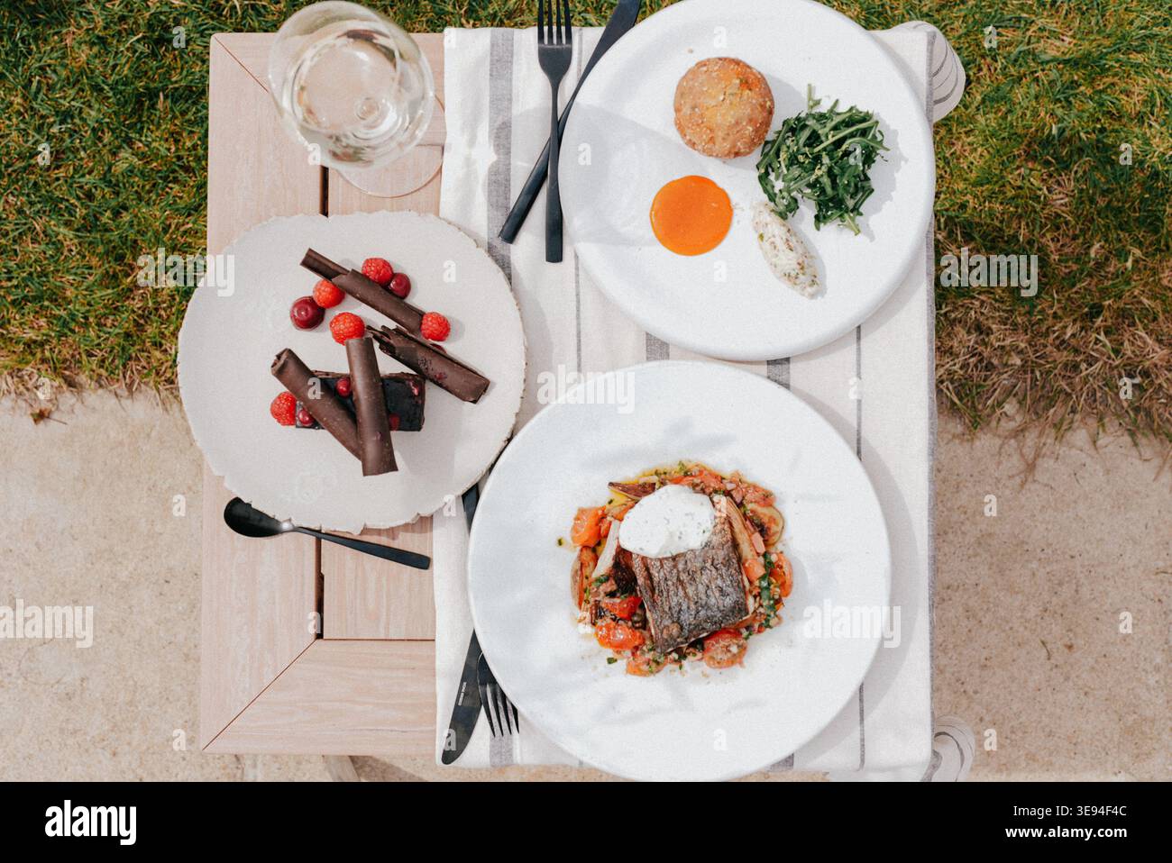 Der Tisch bietet Platz für drei Teller mit frischen, farbenfrohen Speisen. Weiße Teller, Besteck und Gläser ordentlich angeordnet. Gartengras umgibt Holztisch mit Stockfoto