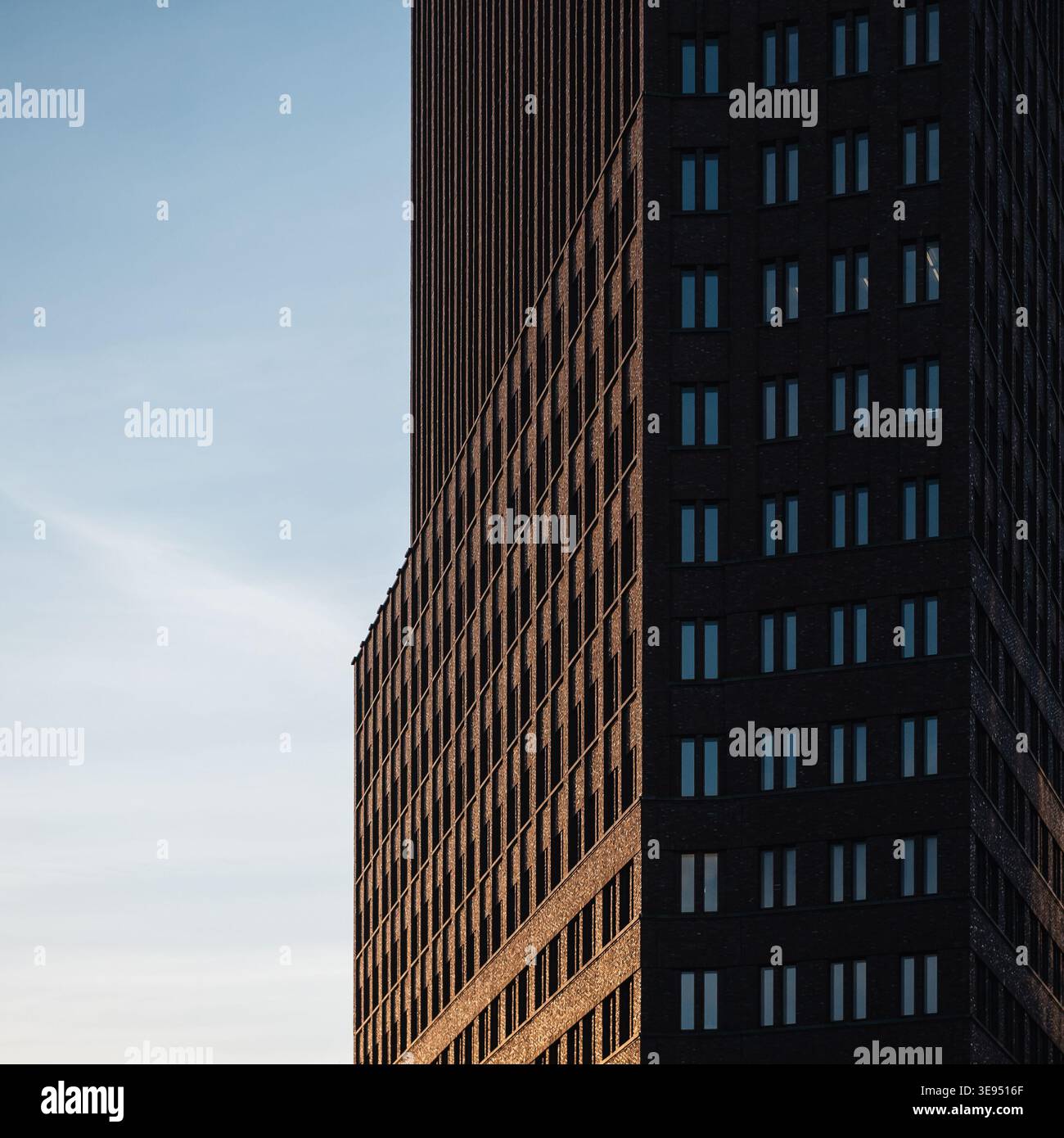 Dunkle Backsteinfassade vor blauem Himmel. Hohes, dunkles Backsteingebäude mit sich wiederholenden Fenstermustern, teilweise von der warmen untergehenden Sonne beleuchtet. Stockfoto