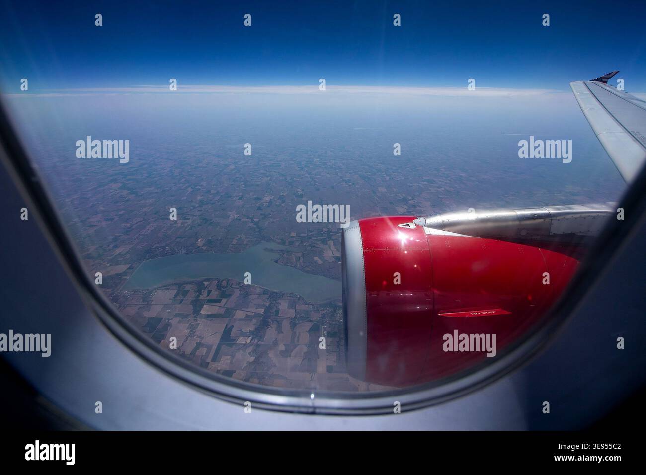 Ein roter Flugzeugmotor ist durch ein kreisförmiges Flugzeugfenster sichtbar, mit einem Flügel nach rechts und einer riesigen Landschaft darunter. Stockfoto