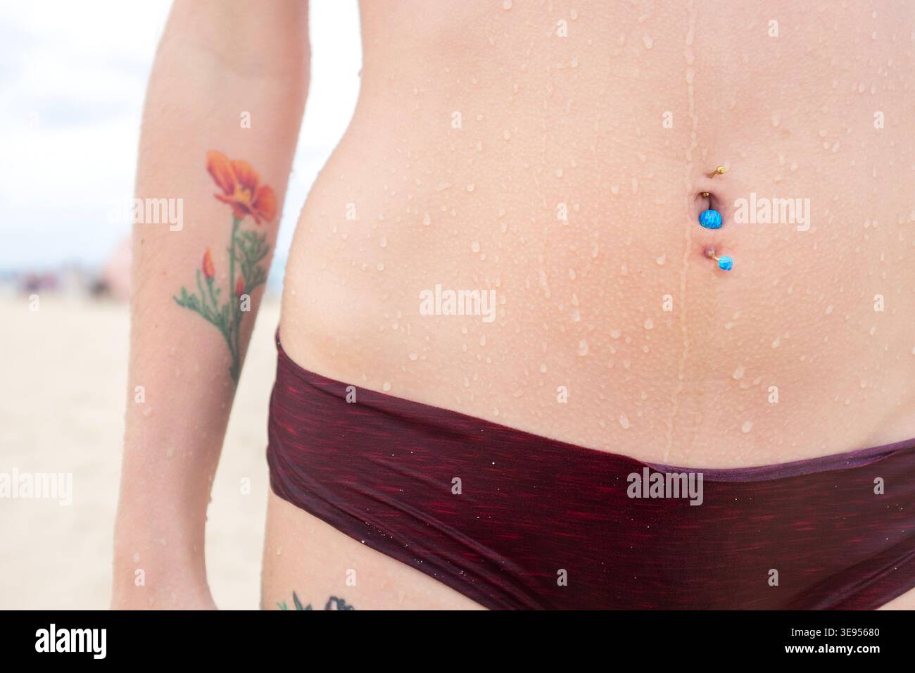 Eine Person steht an einem Sandstrand mit Wassertropfen auf der Haut, trägt eine dunkle Bikinihose und zeigt bunte Piercings auf ihrem Bauchnabel. Stockfoto