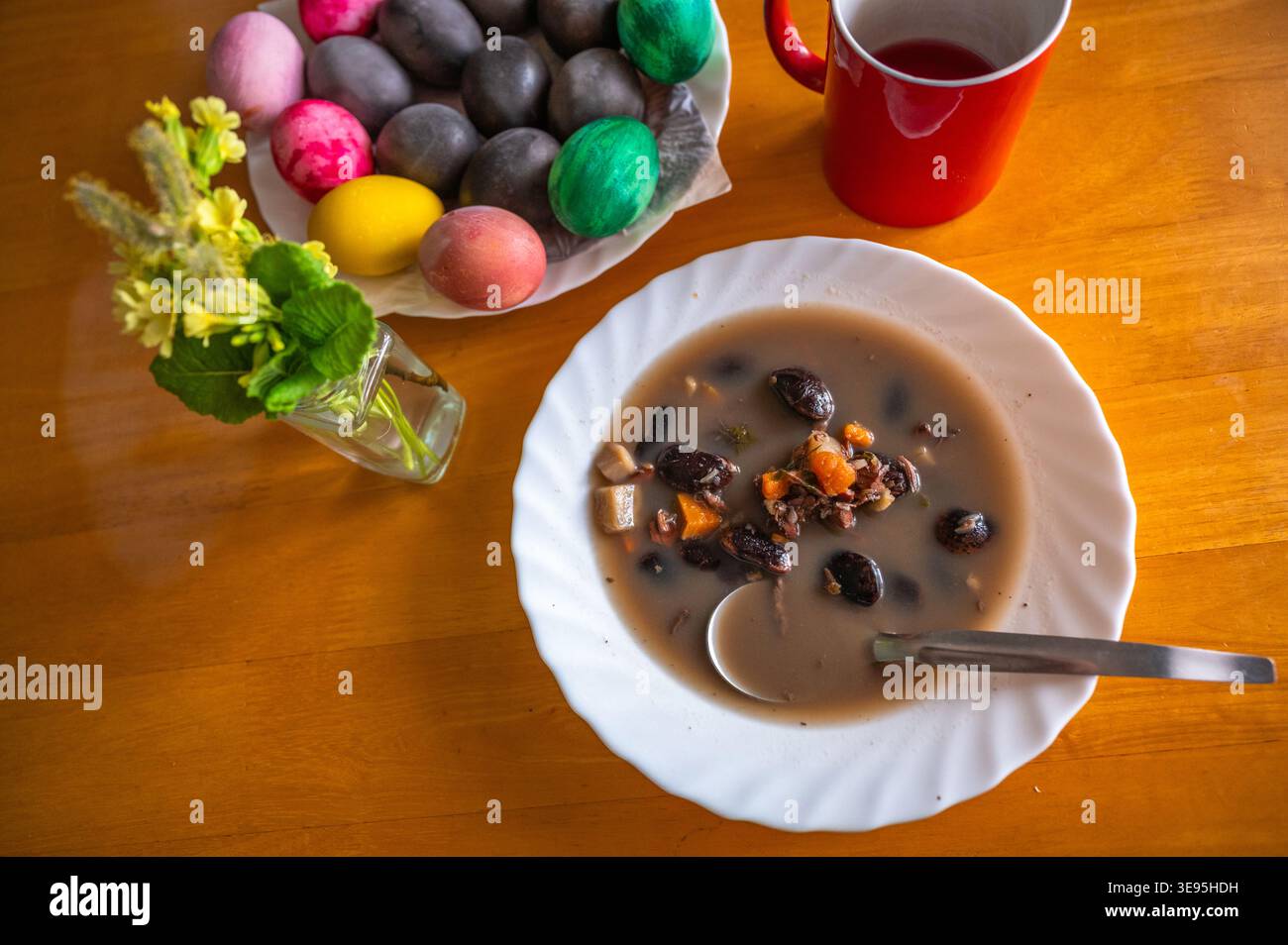 Schwarze Bohnensuppe mit Karotten auf weißem Teller, Löffel, ostereier in Schüssel, Blume in Vase und Teil einer roten Tasse auf dem Tisch. Stockfoto