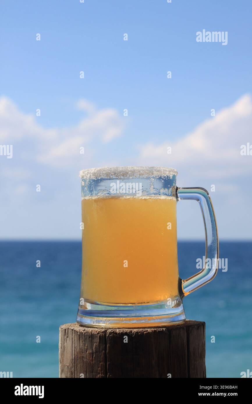 Großes Glas Bier mit gesalzenem Rand vor einem leuchtend blauen Himmel mit weichen weißen Wolken und tiefblauem Meer. Stockfoto