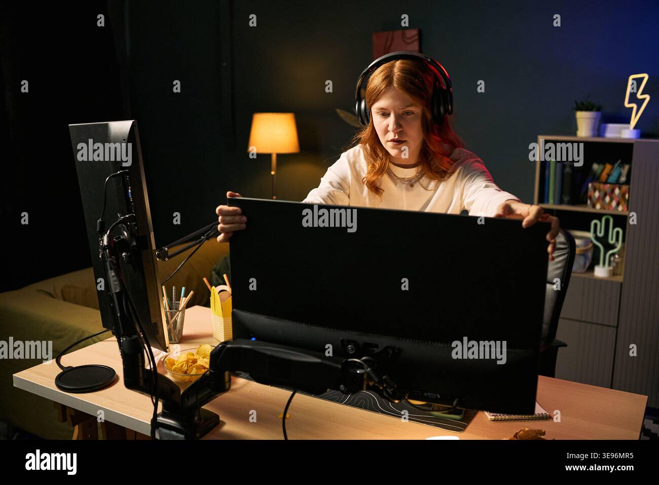 Junge Erwachsene weiße Frau Streamer, der den Monitor anpasst, während er ein Headset am Schreibtisch trägt, um Live-Übertragungen vorzubereiten. Ideal für Gaming-Inhalte, kreative Wirtschaft und Streaming-Plattform-Marketing Stockfoto