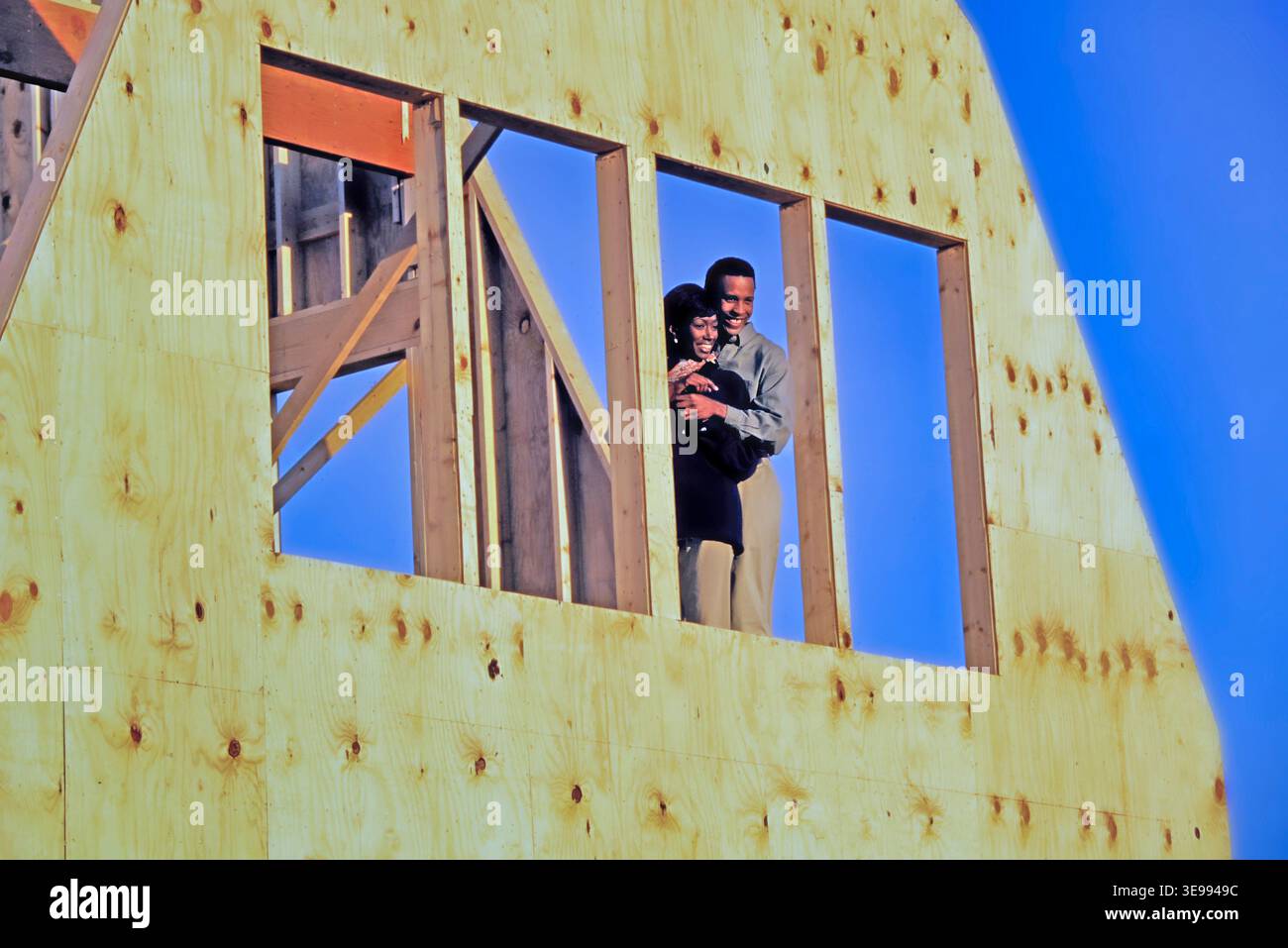 Ein afroamerikanisches Paar lächelt und umarmt sich, während es in einem Fenster ihres neuen Hauses steht, das gerade gebaut wird. Helles Sonnenlicht scheint auf sie im Stockfoto
