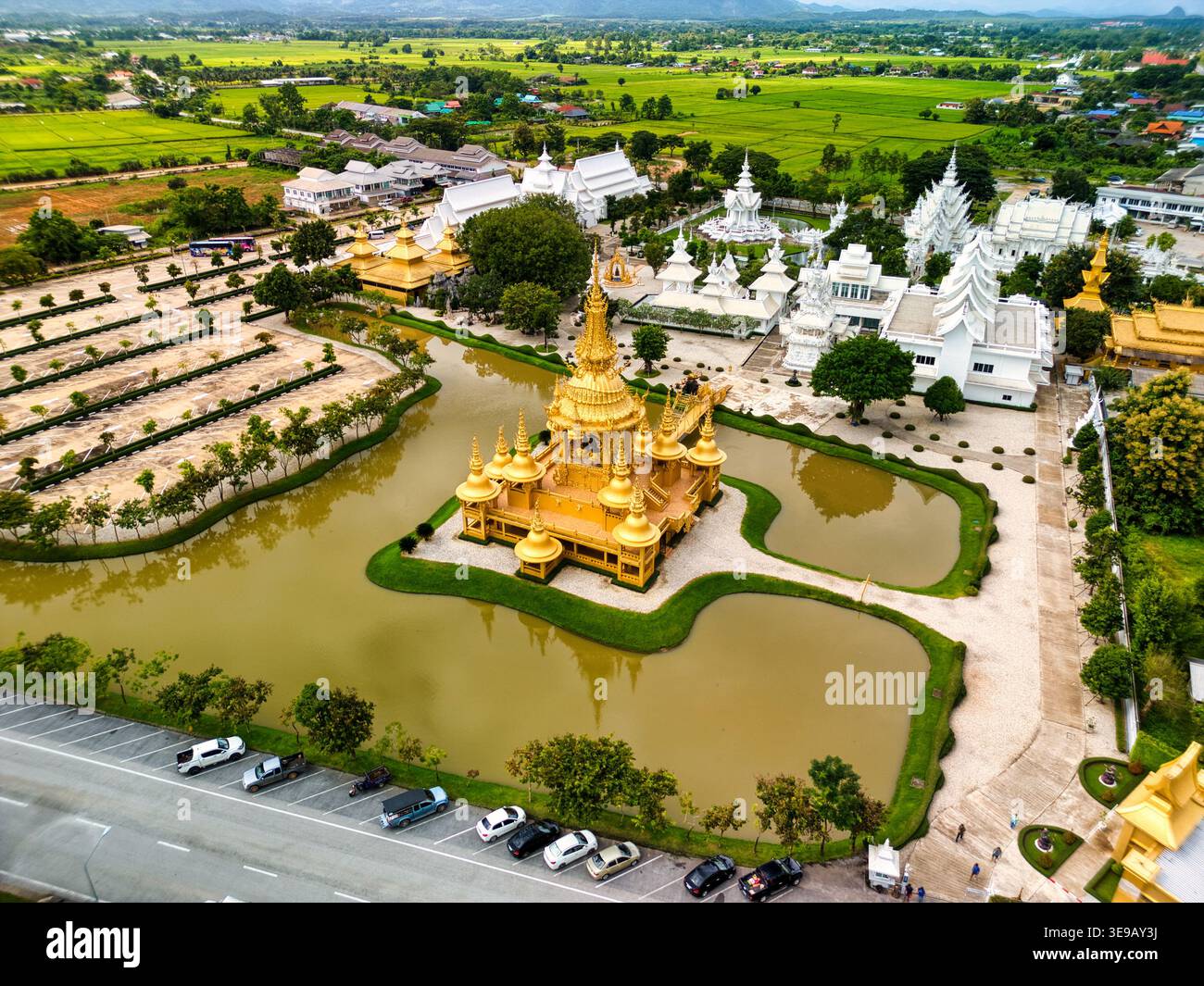 Blick aus der Vogelperspektive auf das kunstvoll verzierte goldene Gebäude am Wat Rong Khun, umgeben von einem reflektierenden Teich und üppigen grünen Feldern in Pa O Don Chai, Chiang Rai, Thailan Stockfoto