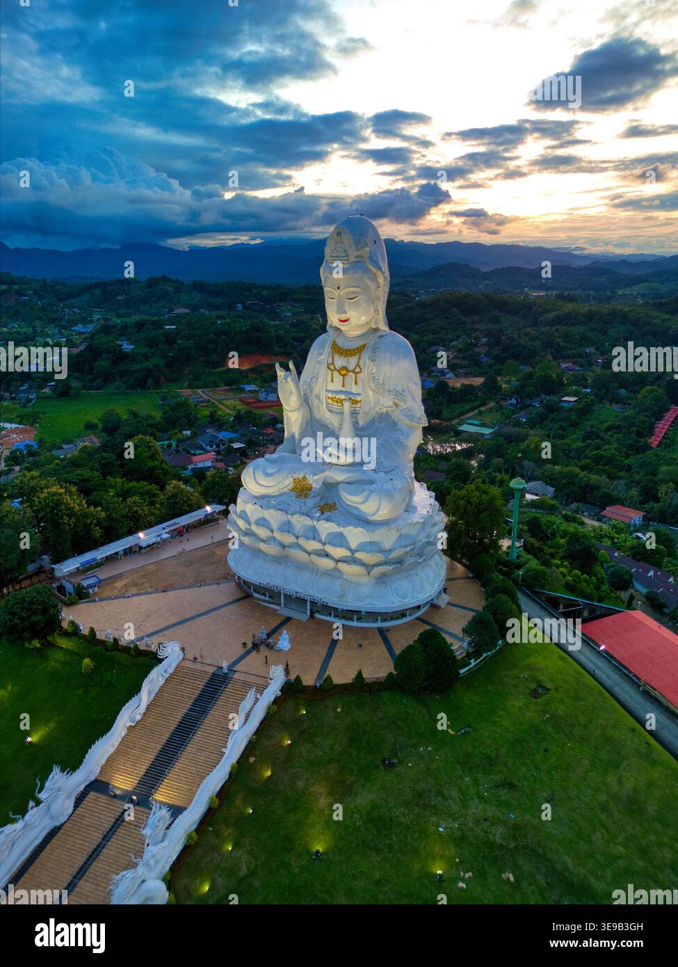 Aus der Vogelperspektive die riesige weiße Guan Yin Statue im Tempelkomplex Wat Huay Pla Kang unter einem dramatischen Sonnenuntergang mit sanften grünen Hügeln in Tambon Ba Stockfoto