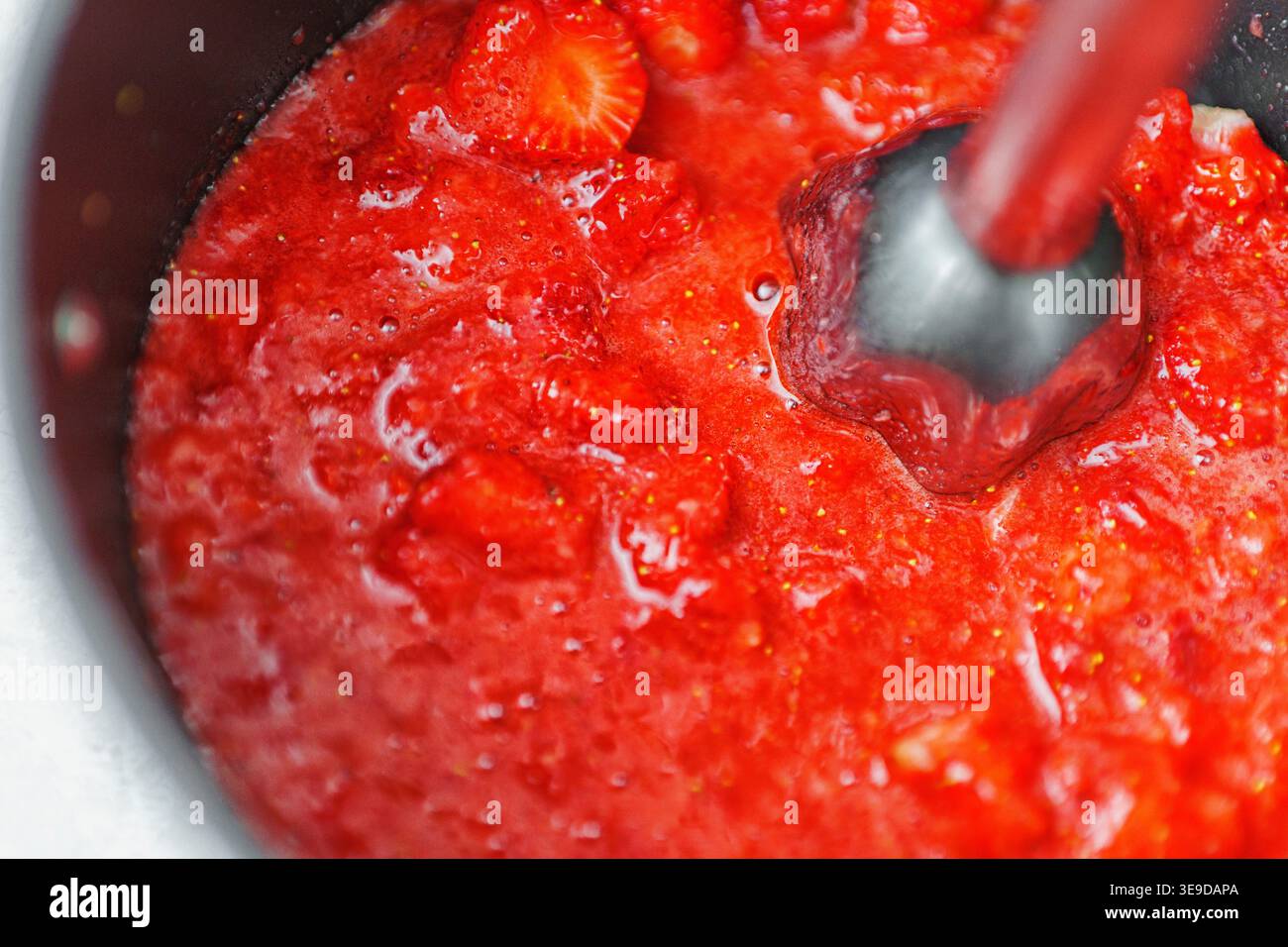 Ein Topf auf einem Herd mit sprudelnder Erdbeermarmelade, die mit einem Löffel gerührt wird. Hausgemachte Erdbeermarmelade. Stockfoto