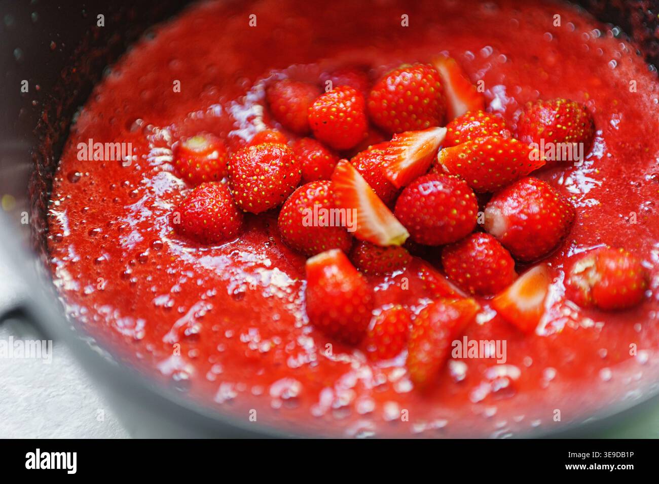 Ein Topf auf einem Herd mit sprudelnder Erdbeermarmelade, die mit einem Löffel gerührt wird. Hausgemachte Erdbeermarmelade. Stockfoto
