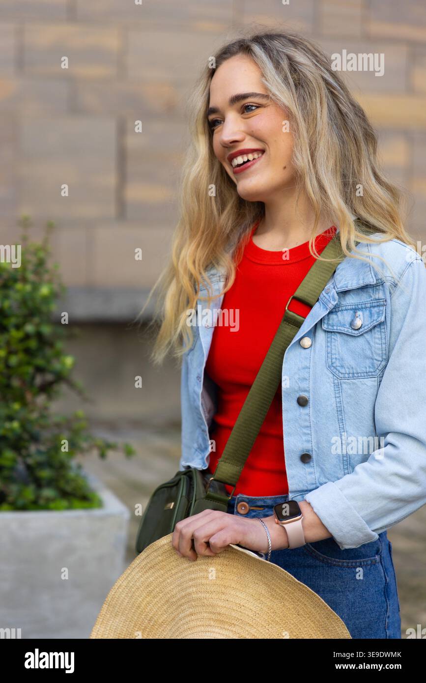 Lächelnde Frau, auf dem platz neben der Steinmauer stehend, mit Strohhut und Jeansjacke. Outdoor, Urban, Freizeit, Crossbody, Uhr, grün, Straßenstil Stockfoto
