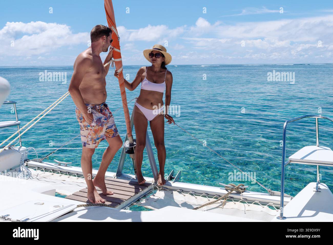 Ein stilvolles Paar entspannt sich auf einem Katamaran auf Mauritius, umgeben von atemberaubendem türkisfarbenem Wasser. Die helle Sonne erleuchtet ihre fröhlichen Ausdrucksformen, während sie den Luxus dieses tropischen Kurzurlaubs genießen. Stockfoto