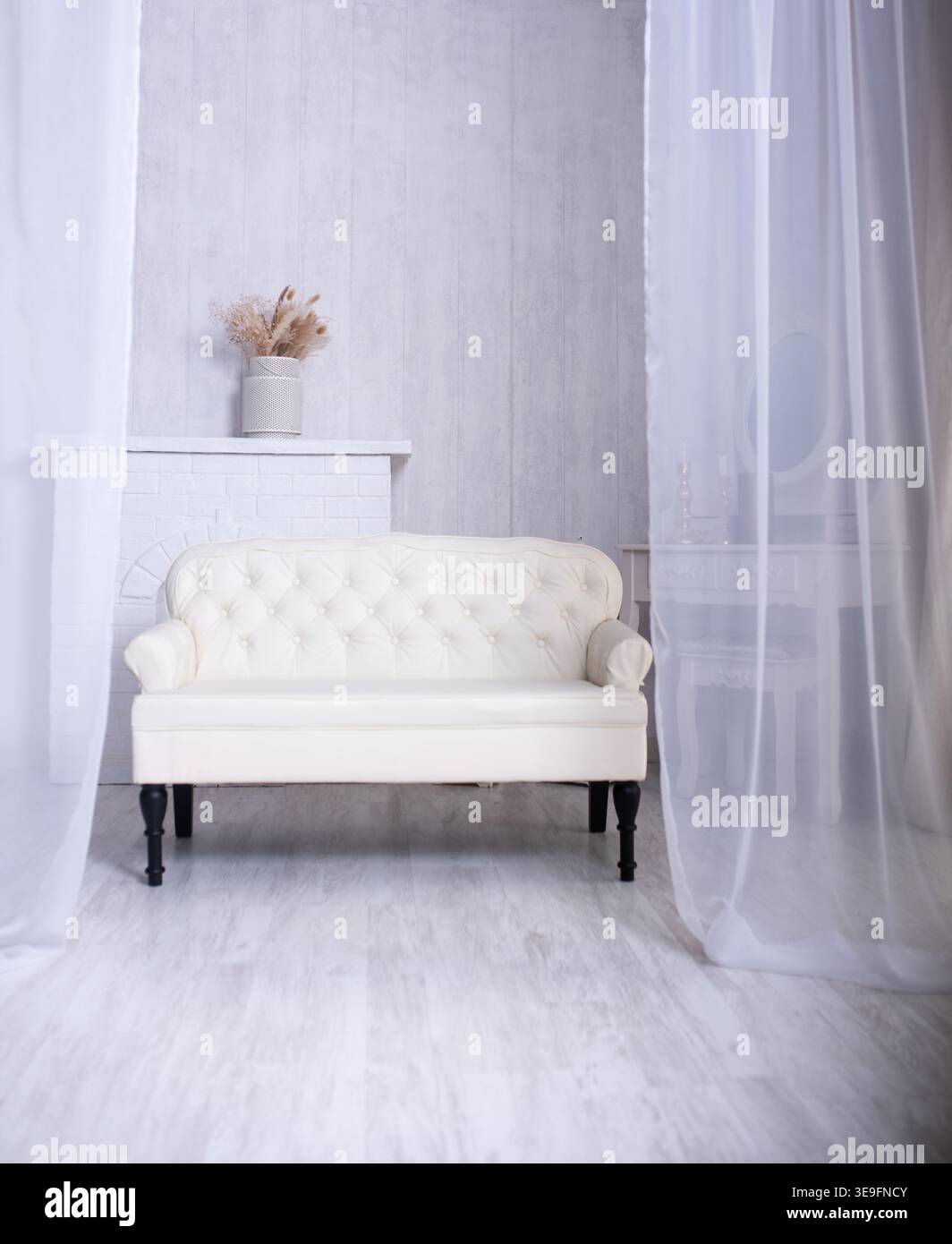 Gemütliches Wohnzimmer, in weißen Tönen dekoriert. Ein klassisches weißes Ledersofa im chesterfield-Stil sitzt vor einem Kaminsims mit getrockneten Elementen Stockfoto