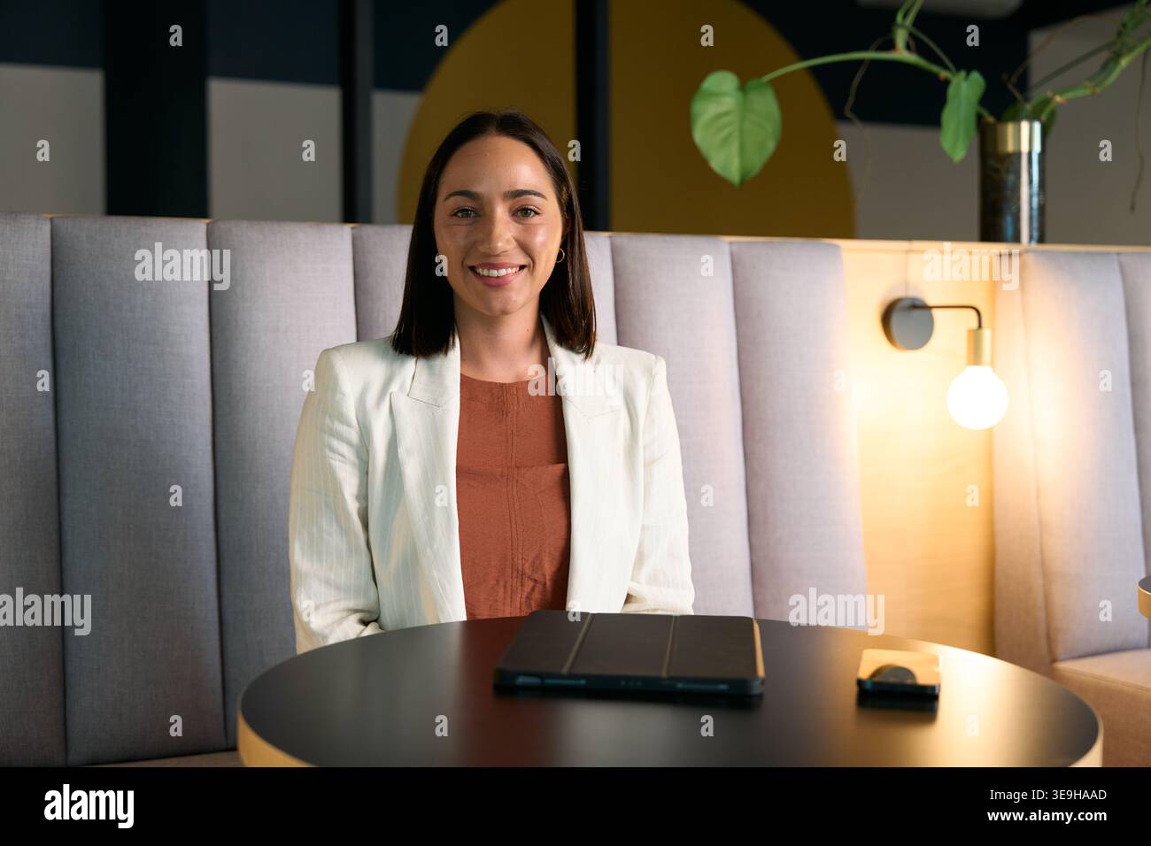Selbstbewusste Geschäftsfrau lächelt, während sie in der Lobby des Büros sitzt und ein digitales Tablet auf dem Tisch liegt Stockfoto