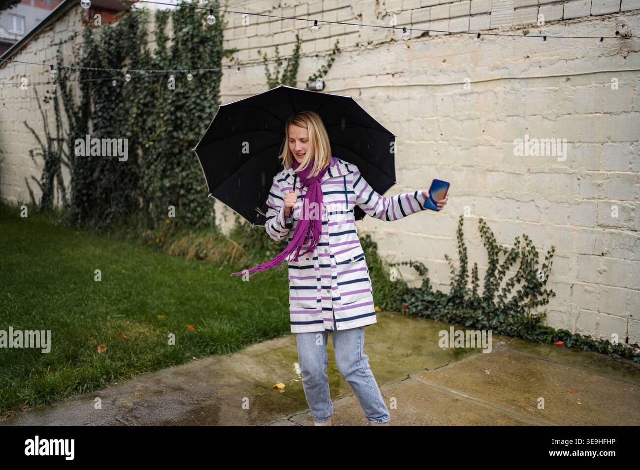 Fröhliche junge Frau, die im Regen tanzt und Schirm und Telefon hält Stockfoto