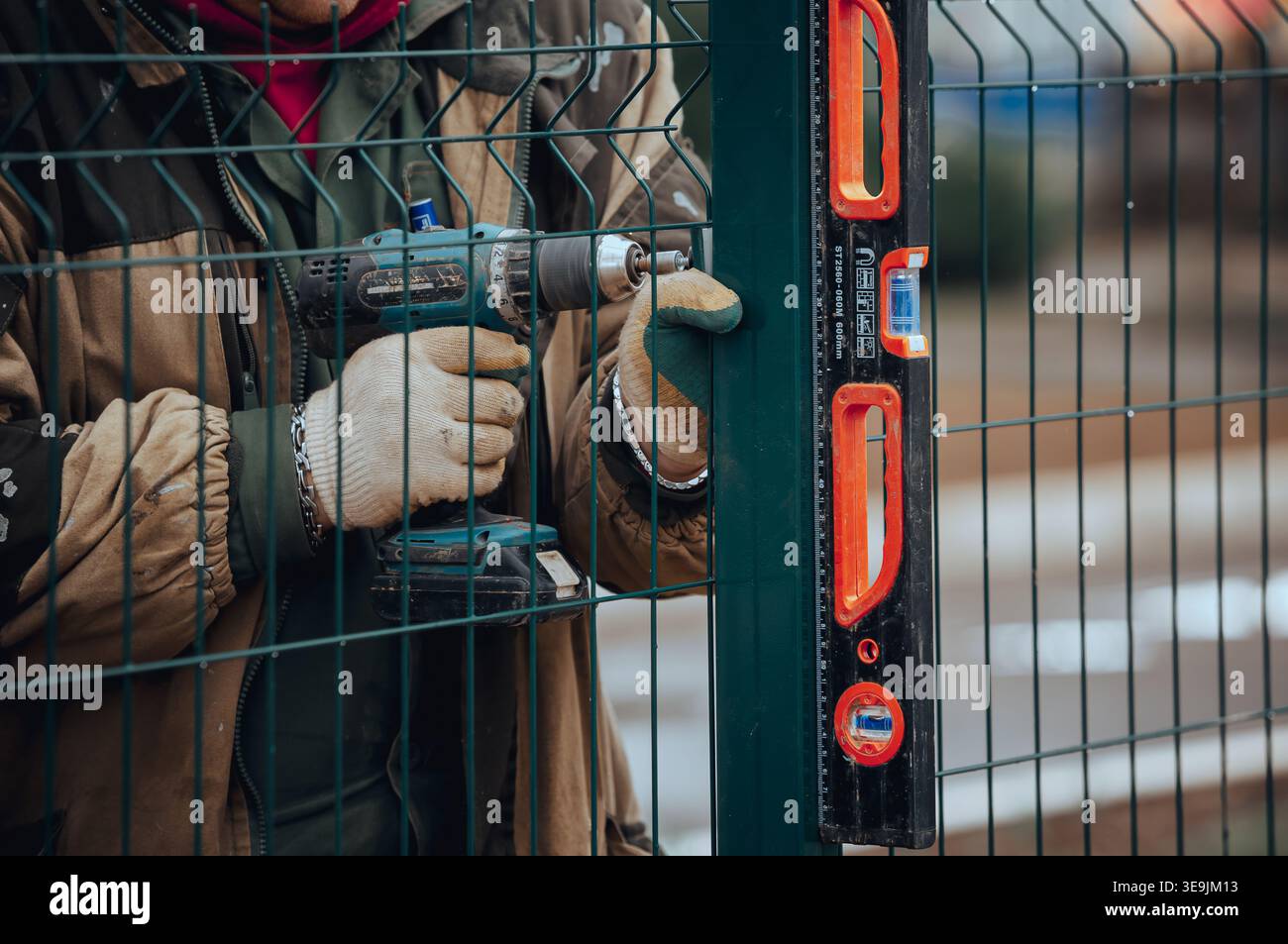 Arbeiter, der einen Metallzaun installiert und die Struktur fixiert, während er die Ausrichtung mit einem waagerechten Werkzeug, manuelle Arbeit im Freien und detaillierte Bauarbeiten überprüft Stockfoto