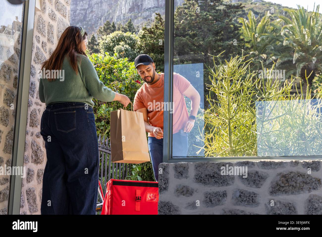 Kurier und Kunde tauschen Papiertüte auf der Veranda aus, Kurier in orangefarbener Kappe mit roter isolierter Tasche Stockfoto