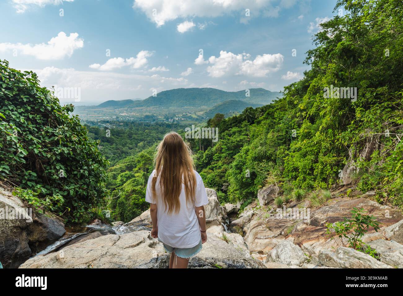 Junge Frau, die auf einer felsigen Klippe mit Blick auf das üppige Dschungeltal und die fernen Hügel in der Nähe des Samui Wasserfalls in Thailand steht Stockfoto
